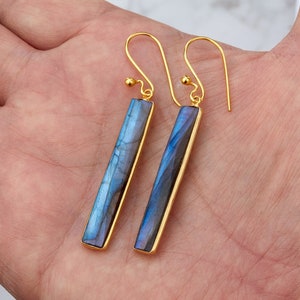 Puede incluir: Un par de pendientes colgantes de oro con colgantes de barra de piedra de labradorita azul. Los pendientes tienen un diseño simple y minimalista.