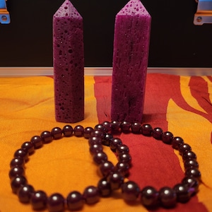 Może przedstawiać: Dwa fioletowe kryształy i sznur ciemnoczerwonych koralików z kamieni szlachetnych. Korale ułożone są w kółko na czerwono-pomarańczowym tle z tkaniny.