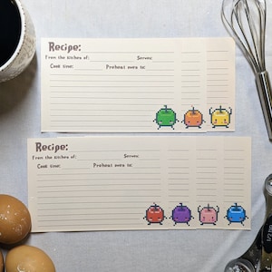 May include: Two blank recipe cards with a pixelated design of colorful characters. The cards have lines for writing down the recipe name, ingredients, instructions, and serving size.
