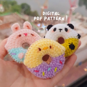 May include: Three crocheted donut plushies, one pink with bunny ears, one yellow with a chick face, and one white with a panda face. The plushies are held in a hand. The text "DIGITAL PDF PATTERN!" is at the top of the image.