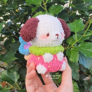 Pu&ograve; includere: Un cane di peluche fatto a mano con pelliccia bianca, orecchie marroni e un corpo rosa e bianco a pois. Il cane ha un colletto verde e un accessorio blu e rosa. Il testo "@windrises. crochet" &egrave; visibile.
