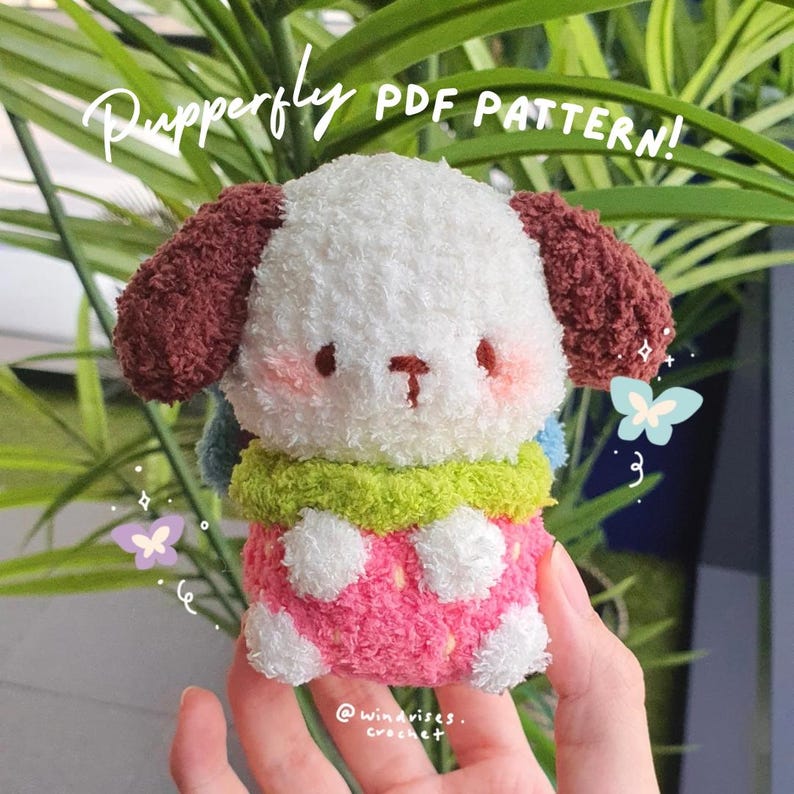 Pu&ograve; includere: Peluche Pupperfly all'uncinetto, con un muso da cane, guance rosa e naso a forma di T. Il peluche &egrave; bianco, rosa, verde e marrone. Il testo "Pupperfly PDF PATTERN!" &egrave; scritto in corsivo.