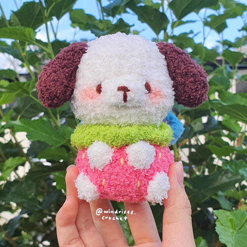 Pu&ograve; includere: Un giocattolo per cani all'uncinetto fatto a mano con un corpo bianco, orecchie bordeaux e una gonna rosa e bianca a pois. Il cane ha un colletto verde e un accento blu. Il giocattolo ha un design carino e da cartone animato. Il testo "@windvises. crochet" &egrave; visibile.