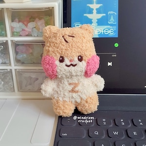 Puede incluir: Un juguete de peluche hecho a mano con forma de oso, con pelaje color melocotón, mejillas rosas y una barriga blanca con la letra "Z". El juguete está sobre un teclado. El texto "@windrises. crochet" es visible.