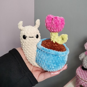 May include: A handmade crochet snail with a blue pot holding a pink flower. The snail is off-white with black eyes and a smiling mouth. The flower has a green stem and a pink bloom. The pot is blue and filled with brown material.