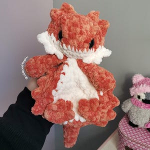 May include: A plush, crocheted lizard toy made of soft, textured orange and white yarn. The creature features a white belly, white spikes along its neck, and dark, round eyes. The text "cuddlecraftbyann" is visible on the side of the plush.