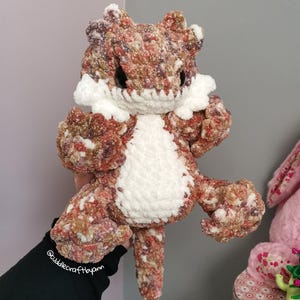 May include: A handmade crochet plush toy of a bearded dragon, crafted from variegated yarn in shades of brown, rust, and cream. The creature features a white textured belly and cheek patches, large black safety eyes, and a long, thick tail.