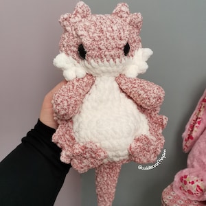 May include: A plush crochet lizard toy featuring a variegated dusty pink and white yarn body with a solid white belly and white cheek tufts. The creature has large black safety eyes and a long tail, with the text @cuddlecraftbyann visible.