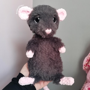 May include: A plush rat toy featuring dark grey fuzzy fur, large black eyes, and light pink accents on the ears, nose, hands, and feet. The soft stuffed animal has a rounded body and a long tail, with the text @cuddlescraftapn visible near the ear.