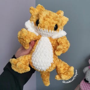 May include: A plush, crocheted bearded dragon toy made from soft, textured golden-yellow yarn with a white belly and white accents around the neck. The creature features large, round, black plastic eyes and a detailed, multi-limbed lizard body.