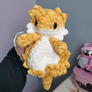May include: A plush, crocheted bearded dragon toy made from soft, textured golden-yellow and white yarn. The stuffed animal features large, round, black plastic eyes, a white belly patch, and a spiky, textured exterior with a long, pointed tail.