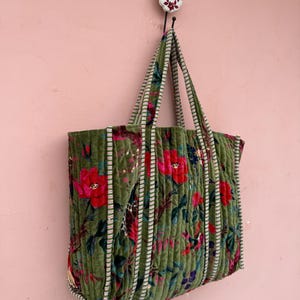 May include: A green velvet tote bag with a floral pattern of red and blue flowers. The bag has striped handles and is hanging on a wall. The bag is made of a soft, quilted material.