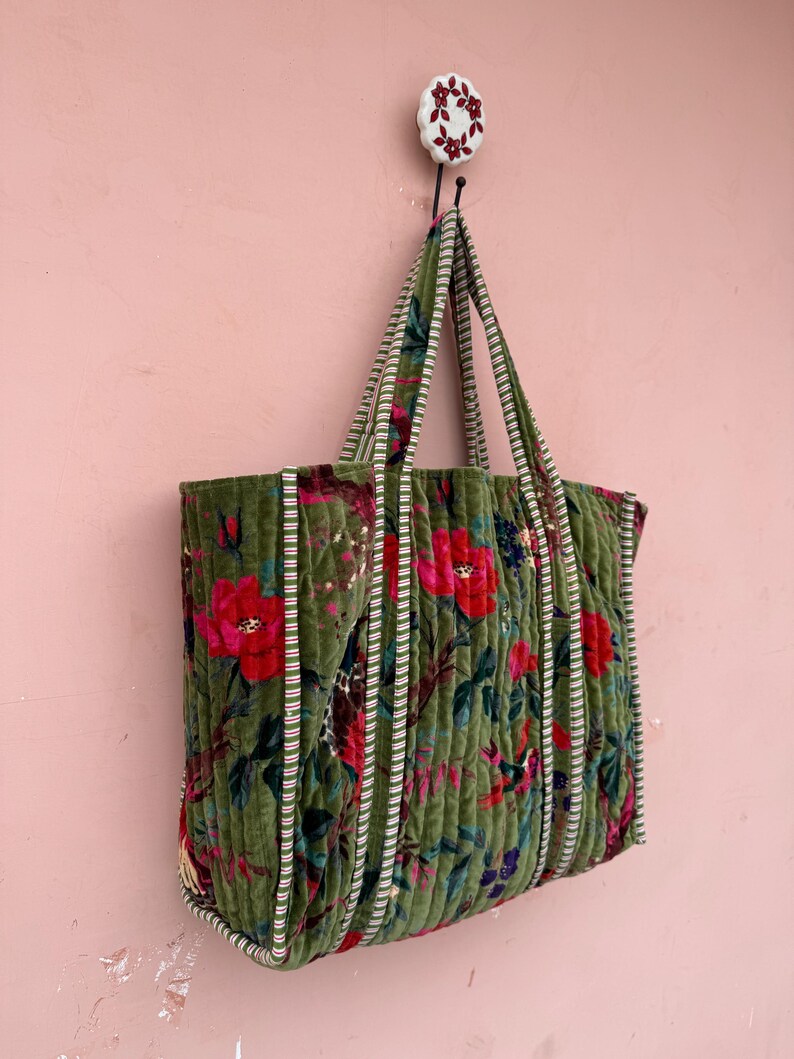 May include: A green velvet tote bag with a floral pattern of red and blue flowers. The bag has striped handles and is hanging on a wall. The bag is made of a soft, quilted material.