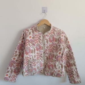 May include: A white quilted jacket with a floral print in shades of pink, orange, and brown. The jacket has a mandarin collar, long sleeves, and two front pockets.