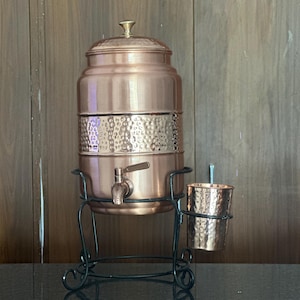 May include: A copper water dispenser with a hammered texture band and a matching copper cup. The dispenser features a metal spigot and a lid with a small knob, all supported by a black metal stand that includes a side holder for the cup.