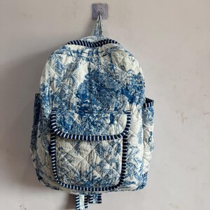 Peut inclure: Sac &agrave; dos matelass&eacute; avec un motif de toile de Jouy bleu et blanc repr&eacute;sentant un paysage. Il poss&egrave;de une poche &agrave; rabat, des poches lat&eacute;rales et une poign&eacute;e sup&eacute;rieure. Les bords sont bord&eacute;s de tissu ray&eacute; bleu et blanc.