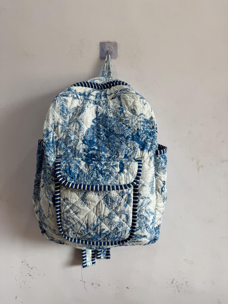 Peut inclure: Sac &agrave; dos matelass&eacute; avec un motif de toile de Jouy bleu et blanc repr&eacute;sentant un paysage. Il poss&egrave;de une poche &agrave; rabat, des poches lat&eacute;rales et une poign&eacute;e sup&eacute;rieure. Les bords sont bord&eacute;s de tissu ray&eacute; bleu et blanc.