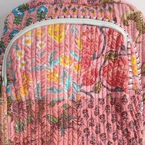 以下が含まれることがあります： ピンク色のキルトバックパック、パッチワークデザイン。バックパックは、ピンク、黄色、青、赤の色合いの花柄と鳥柄が特徴です。白いジッパーと上部のハンドルが付いています。