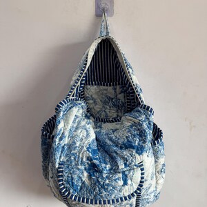 Peut inclure: Un sac fourre-tout matelass&eacute; avec un motif de toile de Jouy bleu et blanc, repr&eacute;sentant un paysage. Le sac a un int&eacute;rieur et une bordure ray&eacute;s, avec une poign&eacute;e. Le sac est accroch&eacute; au mur.
