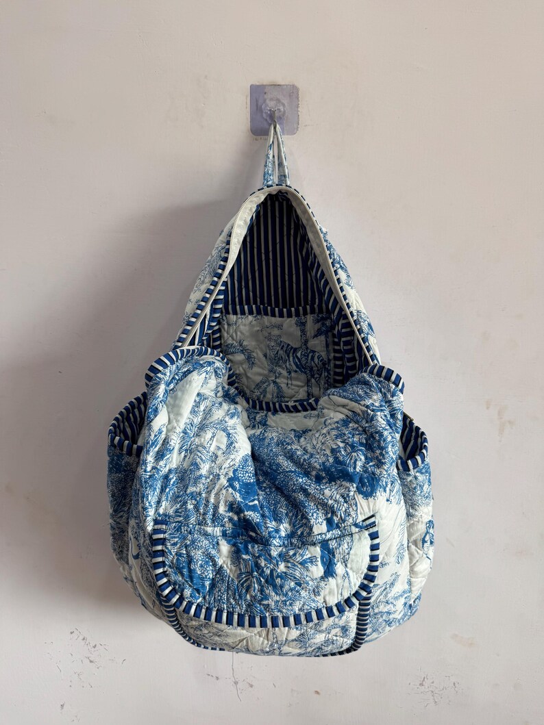 Peut inclure: Un sac fourre-tout matelass&eacute; avec un motif de toile de Jouy bleu et blanc, repr&eacute;sentant un paysage. Le sac a un int&eacute;rieur et une bordure ray&eacute;s, avec une poign&eacute;e. Le sac est accroch&eacute; au mur.