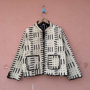 Peut inclure: Veste crème avec un motif géométrique noir. La veste a un col mandarin, deux poches avant et une fermeture éclair sur toute la longueur. Le design présente des lignes et des points verticaux et horizontaux.