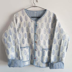 May include: A white quilted jacket with a blue floral print and blue and white striped trim. The jacket has a button closure and two pockets.