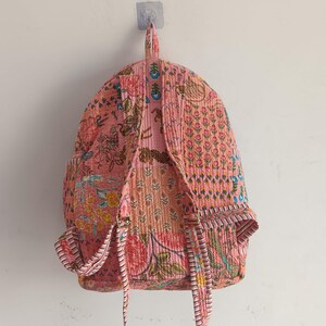 以下が含まれることがあります： ピンク色のキルトバックパック、花柄。 バックパックには調節可能なストラップ、上部のハンドル、フロントポケットが付いています。 生地は、ピンク、オレンジ、緑の色合いのさまざまな花柄のデザインのパッチワークです。