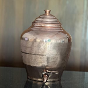 Pode incluir: Um dispensador de água de cobre polido com corpo arredondado, tampa escalonada e uma pequena torneira de metal na base. A seção central apresenta uma textura de faixas horizontais escovadas, enquanto as partes superior e inferior são lisas.