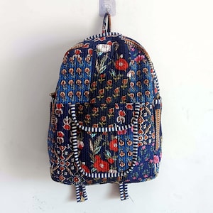 May include: A quilted backpack featuring a floral design in blue, red, and white. The bag has a front pocket with a striped border and adjustable straps. The backpack is hanging on a white wall.