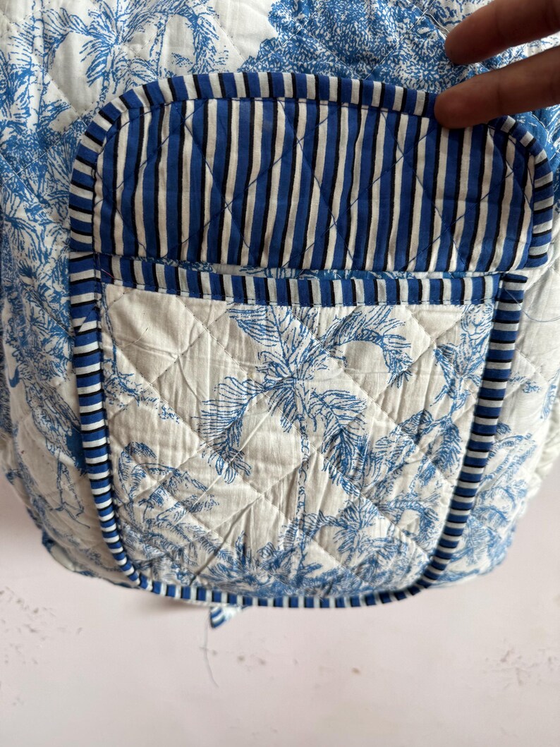 Peut inclure: Tissu matelass&eacute; avec un motif de palmiers tropicaux bleus et blancs. La poche pr&eacute;sente un motif &agrave; rayures bleues et blanches. Les bords sont bord&eacute;s d'une bordure ray&eacute;e bleue et blanche. Le tissu est matelass&eacute; avec un motif en losange.