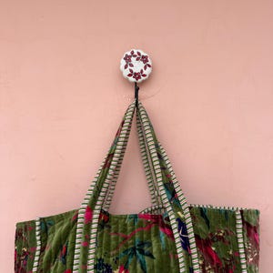 May include: A green floral tote bag with striped handles hangs from a white and red floral hook against a pink wall. The bag features a quilted design with red and pink flowers and green leaves.