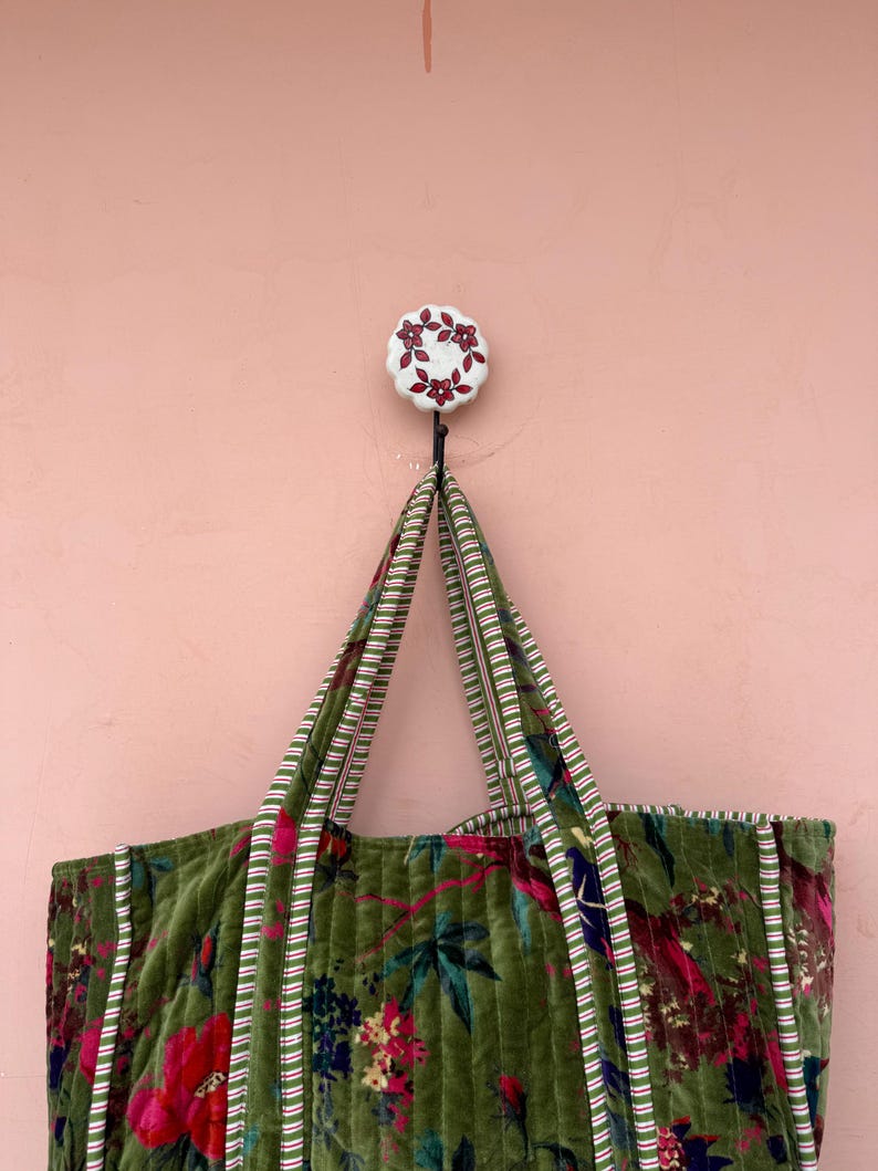 May include: A green floral tote bag with striped handles hangs from a white and red floral hook against a pink wall. The bag features a quilted design with red and pink flowers and green leaves.