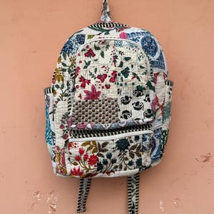 Puede incluir: Una mochila acolchada con un diseño de patchwork con estampados florales en tonos rojos, rosas, azules y verdes sobre un fondo blanco. La mochila tiene dos bolsillos laterales y correas a rayas.