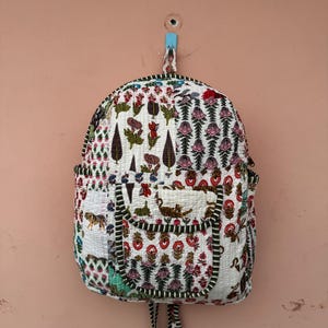 Op de afbeelding: Een witte gewatteerde rugzak met een kleurrijke bloemen- en dierenprint. De rugzak heeft een voorvak, zijvakken en een zwart-witte gestreepte rand. Hij hangt aan een roze muur.