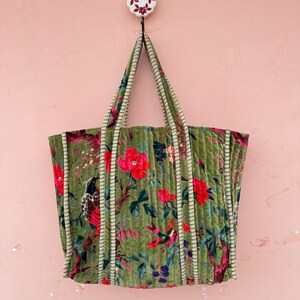 May include: A green tote bag with a floral pattern of red and pink flowers, birds, and foliage. The bag has striped handles and trim. The bag is hanging on a white hook with a floral design against a pink wall.