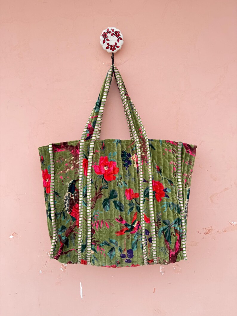 May include: A green tote bag with a floral pattern of red and pink flowers, birds, and foliage. The bag has striped handles and trim. The bag is hanging on a white hook with a floral design against a pink wall.