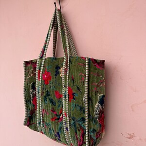 May include: A green quilted tote bag with a floral and bird pattern. The bag has striped handles and trim. The bag is hanging on a decorative hook against a pink wall.