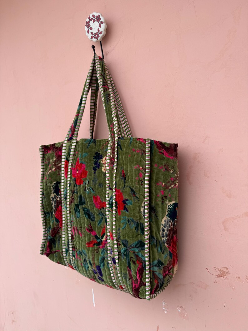 May include: A green quilted tote bag with a floral and bird pattern. The bag has striped handles and trim. The bag is hanging on a decorative hook against a pink wall.