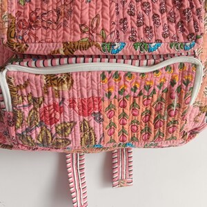 以下が含まれることがあります： ピンク色のキルトバックパック。花柄と鳥柄が特徴です。白いトリムとストライプのストラップが付いたジッパーポケットがあります。生地は垂直方向にステッチされています。