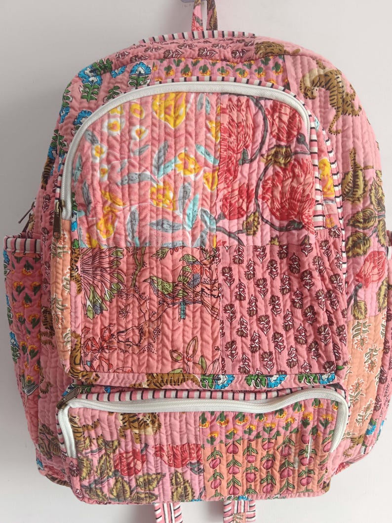 以下が含まれることがあります： ピンク色のキルトバックパック。花柄とペイズリー柄が特徴です。フロントジッパーポケット、サイドポケット、白いジッパーが付いています。トップハンドルと調節可能なストラップ付きです。