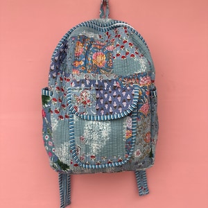 Puede incluir: Una mochila acolchada en tonos azules, con estampados florales y paisley. La bolsa tiene un cierre de solapa, bolsillos laterales y correas ajustables. La mochila está colgada contra una pared rosa.