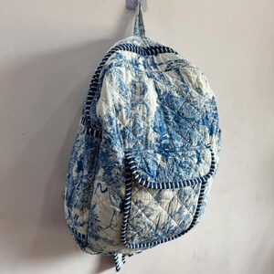 Peut inclure: Sac &agrave; dos matelass&eacute; avec un imprim&eacute; tropical bleu et blanc. Le sac &agrave; dos est dot&eacute; d'une poche avant et est bord&eacute; d'un passepoil bleu. Le sac est accroch&eacute; &agrave; un mur.