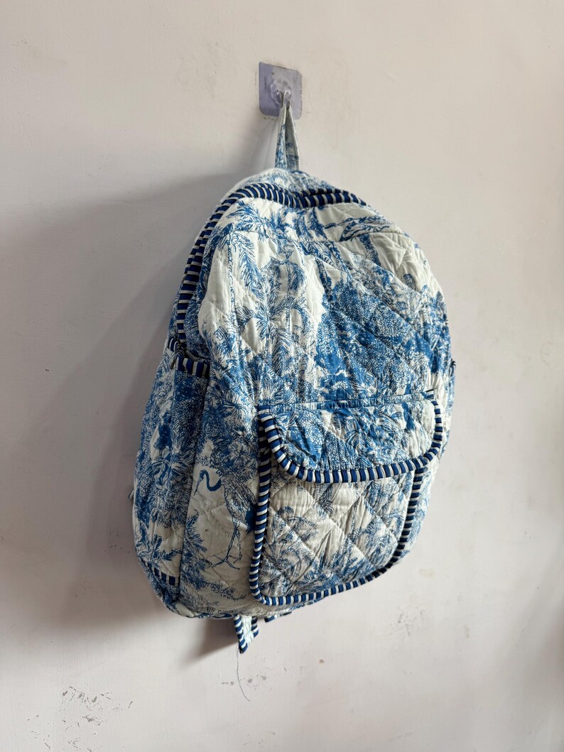 Peut inclure: Sac &agrave; dos matelass&eacute; avec un imprim&eacute; tropical bleu et blanc. Le sac &agrave; dos est dot&eacute; d'une poche avant et est bord&eacute; d'un passepoil bleu. Le sac est accroch&eacute; &agrave; un mur.