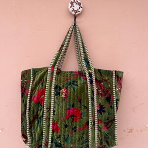 May include: A large, olive green tote bag with a floral pattern of red and pink flowers. The bag has striped trim and handles, and is hanging from a decorative wall hook. The bag is made of a quilted velvet material.