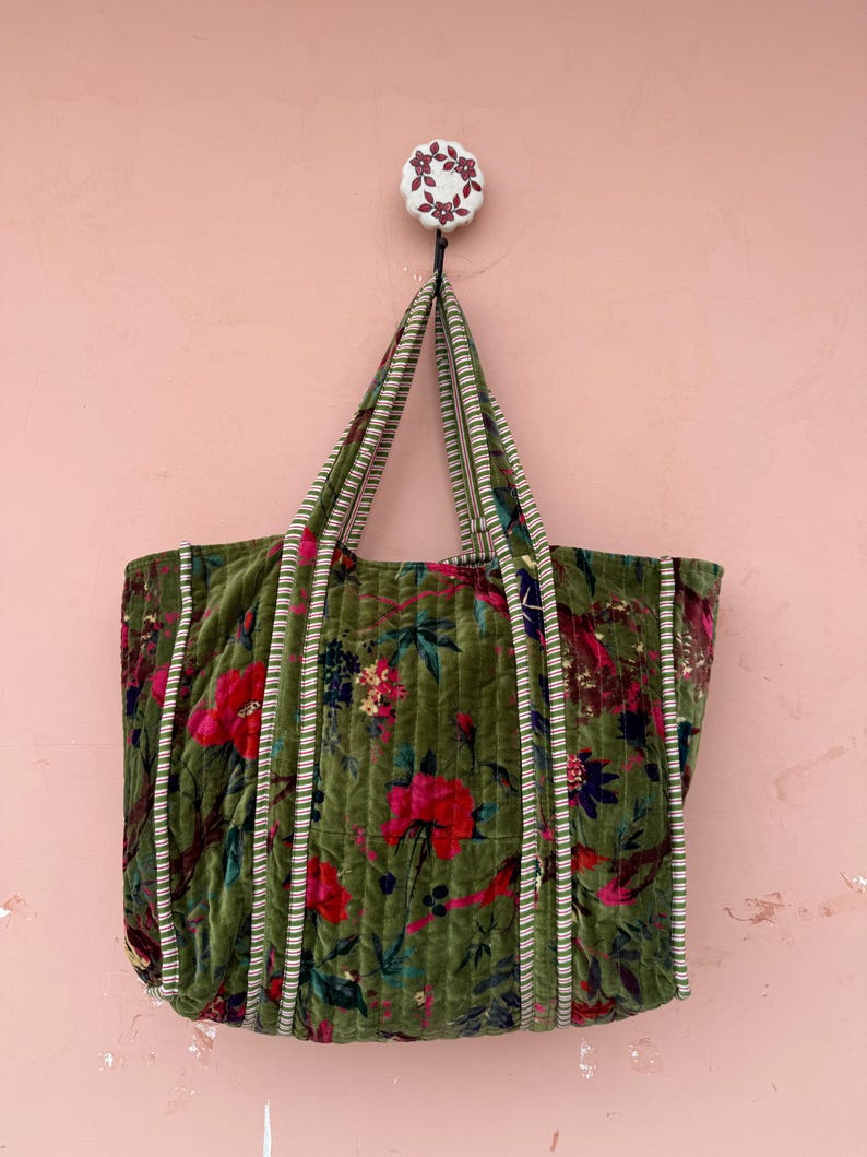 May include: A large, olive green tote bag with a floral pattern of red and pink flowers. The bag has striped trim and handles, and is hanging from a decorative wall hook. The bag is made of a quilted velvet material.