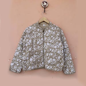 Peut inclure: Veste matelassée à motifs floraux blancs et vert olive. La veste a deux poches avant et un col rayé. Les bords sont bordés d'une matière foncée ressemblant à une corde. Elle est suspendue à un cintre en bois.