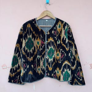 May include: A black velvet jacket with a colorful ikat pattern in green, gold, and red. The jacket has a round neckline, long sleeves, and a button-up front. The lining is striped in black and white.