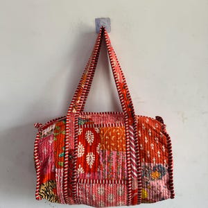 Op de afbeelding: Een gewatteerde tote bag met een patchwork ontwerp in tinten rood, roze en oranje. De tas heeft een rechthoekige vorm met twee handvatten en is afgewerkt met rood-wit gestreepte stof. De tas hangt aan een witte muur.