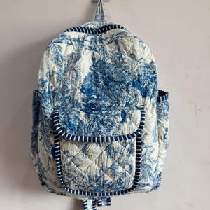 Peut inclure: Un sac &agrave; dos matelass&eacute; avec un motif de toile de Jouy bleu et blanc. Il poss&egrave;de une poche &agrave; rabat, des poches lat&eacute;rales et des bordures bleues. Le sac est accroch&eacute; au mur.