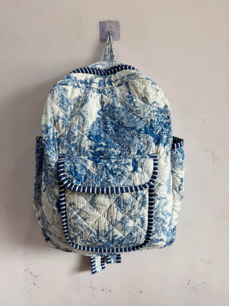 Peut inclure: Un sac &agrave; dos matelass&eacute; avec un motif de toile de Jouy bleu et blanc. Il poss&egrave;de une poche &agrave; rabat, des poches lat&eacute;rales et des bordures bleues. Le sac est accroch&eacute; au mur.